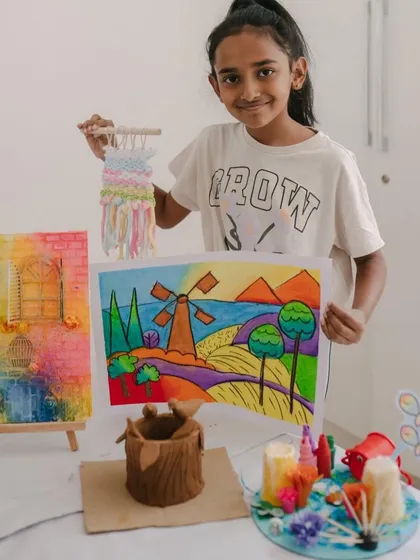 This young artist is proudly presenting her portfolio of work from our summer art camp. Her collection includes a vibrant landscape painting, a small woven piece, a sculpted pot, and a 3D diorama, showcasing the wide range of skills she explored.