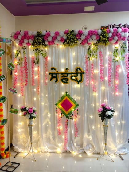 This simple and sober Mehndi function decor is perfect for intimate home events. It features a white backdrop with fairy lights, a pink and white floral header, and hanging pink flowers.