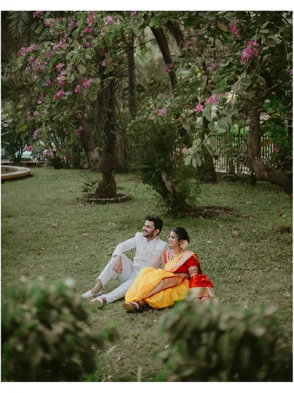 A wide shot of the couple sharing a relaxed moment on the grass in a beautiful park, surrounded by flowering trees.