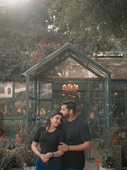 A beautiful, sun-drenched pre-wedding photo of a couple in a garden. The groom kisses the bride's forehead under a canopy of trees, creating a fairytale-like moment.