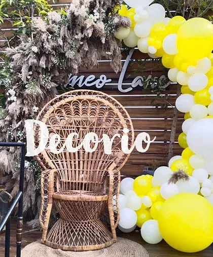 This boho-chic baby shower setup features a wicker peacock chair as the main seat for the mom-to-be. The scene is framed by a yellow and white balloon arch and dried pampas grass for a natural, trendy look.