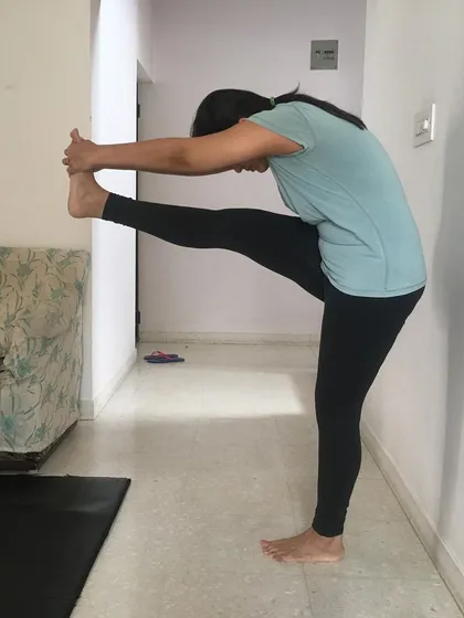 A student practices Dandayamana Janushirasana (Standing Head-to-Knee Pose) with wall support. In online classes, we use simple props available at home to make poses more accessible.