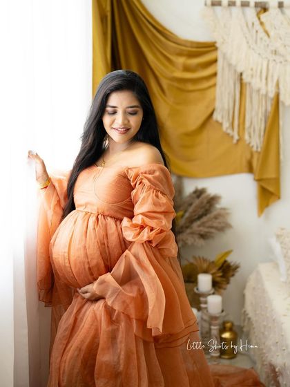 A glowing mother-to-be stands by a window in a beautiful peach gown. The soft light illuminates her in this warm, boho-styled room, creating a peaceful and radiant portrait.