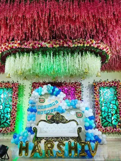 This grand stage was designed for a 'Welcome Baby' ceremony, featuring an ornate white throne-style sofa for the family. The backdrop includes a dense balloon arch in shades of blue and white, custom banners, and large gold foil balloons spelling out the baby's name, all under an elaborate ceiling of hanging flowers.