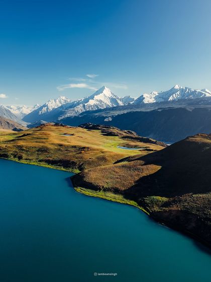The breathtaking Chandratal Lake, also known as the Moon Lake, with snow-dusted peaks in the background. This drone shot captures the vibrant turquoise water against the barren landscape of Spiti.