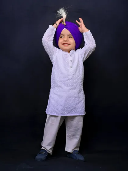 A young Sikh boy in a white kurta and a vibrant purple turban smiles joyfully. This individual portrait, part of a family session, captures his bright and happy personality.