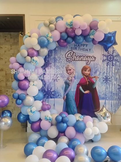 A magical Frozen themed birthday party featuring a circular backdrop with Elsa and Anna, surrounded by a blue, purple, and white balloon garland.
