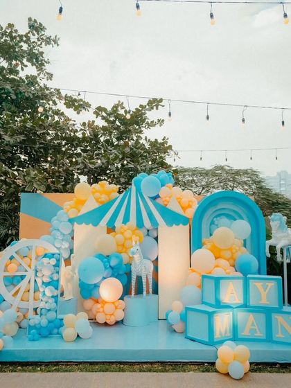 A wide shot of the blue and yellow carnival-themed first birthday stage. The setup includes a carousel horse, a tent, and a large number "1" prop filled with balloons.