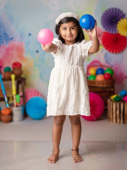 Happy Holi! This little girl is ready to play with colors, holding up pink and blue balls with a joyful smile.