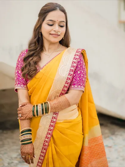 A full-length view of a simple yet stunning Marathi bridal look. The bride's yellow saree with a contrasting pink blouse is complemented by minimal makeup, soft waves, and traditional green bangles.