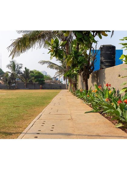 A paved walkway runs alongside the main lawn, bordered by hardy, flowering plants. This provides a clear circulation path for walkers and joggers while separating them from the central play area at the Worli ground.