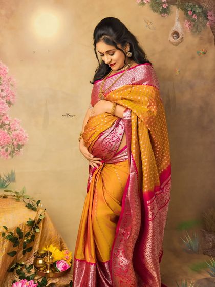 A standing solo portrait capturing the radiant glow of pregnancy. The rich silk saree drapes beautifully over her baby bump, creating a timeless and elegant look.
