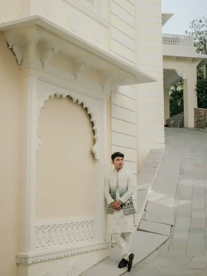 A solo portrait of the groom looking sharp in his traditional attire against the pristine white architecture of a Udaipur palace. It's a moment of quiet confidence before the big day.