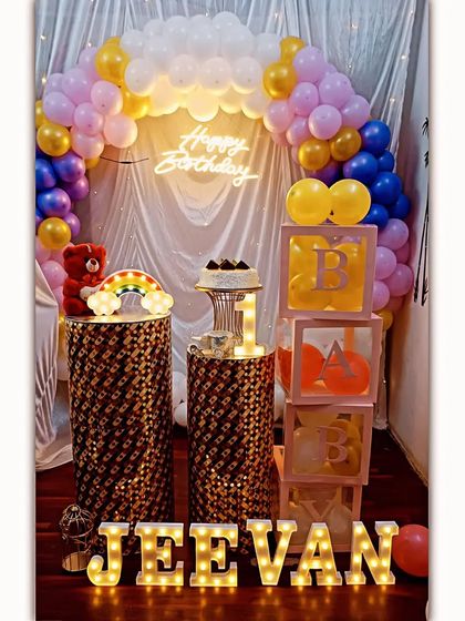 A first birthday decoration with a balloon arch, a neon sign, and 'BABY' display boxes. The sequined pedestals and light-up marquee letters add a touch of sparkle.