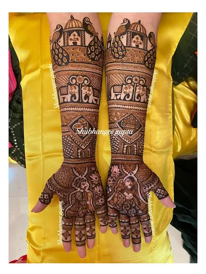 A beautiful and happy bride showing off her full wedding mehndi. The design includes portraits, elephants, and peacocks, covering her hands and arms in traditional wedding symbols.