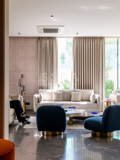 Another angle of the 'House Retold' living room, showing the interplay of textures between the panelled wall, plush sofas, and smooth marble floor. The deep blue chairs provide a strong color contrast.
