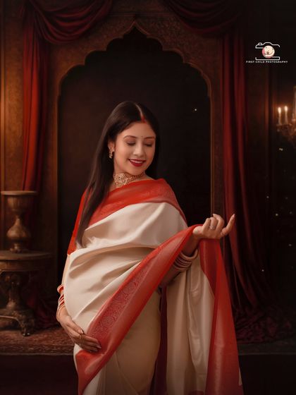 A close-up shot focusing on the gentle expression of the expecting mother. Her smile and downward glance towards her bump convey a deep sense of love, with the rich red and cream saree framing the moment.
