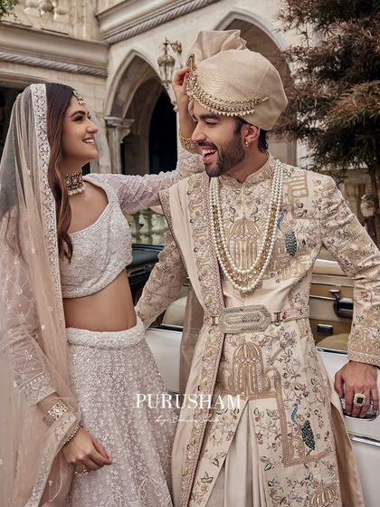 A playful, candid moment. The groom's sherwani, with its unique peacock and architectural embroidery, is a piece of wearable art, accessorized with a pearl necklace and a custom belt.