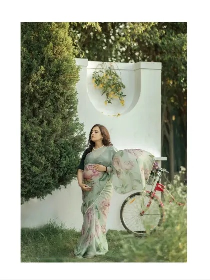 A full-length portrait showcasing the beautiful drape of the floral saree in a lovely garden setting. The mother-to-be's gentle hold on her belly speaks of love and protection.