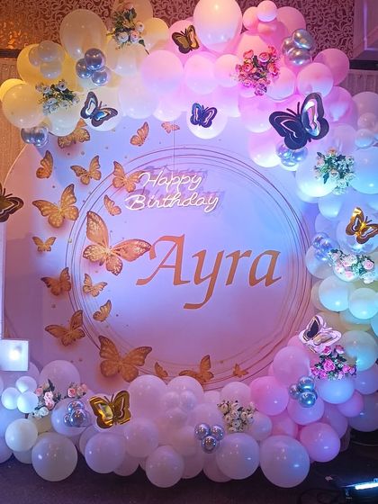 A whimsical butterfly theme for a first birthday party. The circular backdrop is adorned with golden butterflies and a pastel balloon garland, complemented by a light-up number '1'.