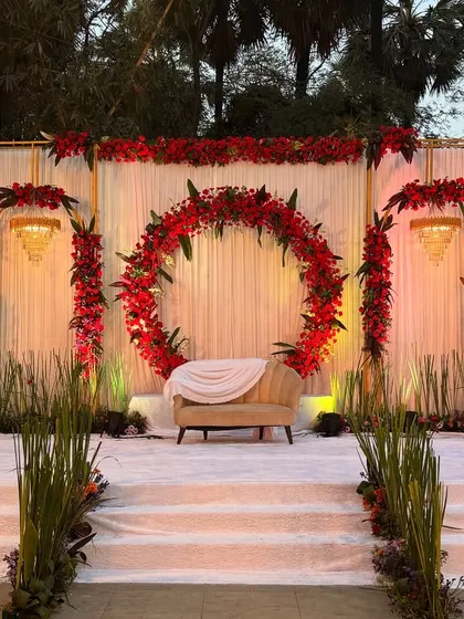 A beautiful reception stage with a red and white theme. The central floral ring and hanging chandeliers create an elegant focal point for the couple's seating area.