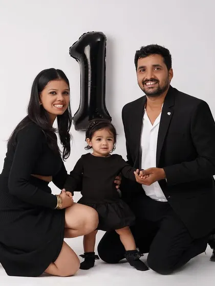 A happy family of three kneels together for a first birthday photo, smiling at the camera with a '1' balloon in the background.