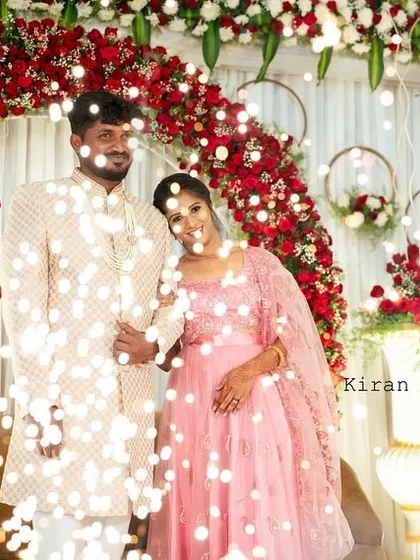 An happy couple at their engagement, framed by a beautiful arch of red and white roses. The fairy lights and hanging floral hoops in the background create a whimsical and romantic setting.