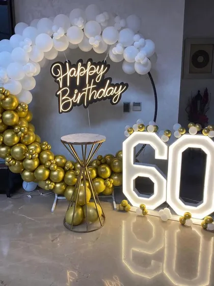 An elegant 60th birthday party backdrop in gold and white. This setup combines a circular balloon arch, a stylish 'Happy Birthday' neon sign, and large marquee numbers to celebrate the milestone.