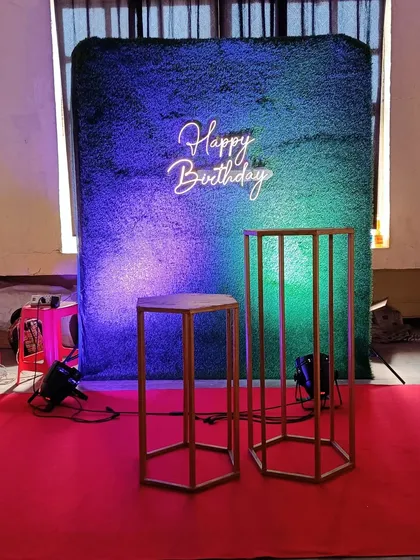 A minimalist yet effective birthday setup, featuring a green turf wall, a 'Happy Birthday' neon sign, and display pedestals, ready for a cake or other decor.