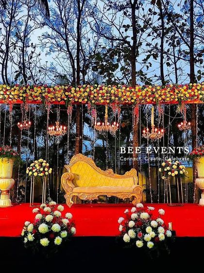 An outdoor reception setup against a backdrop of trees. The stage is framed with a beautiful arch of mixed flowers, with hanging chandeliers and a classic golden chaise lounge, blending natural beauty with classic elegance.