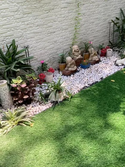 This garden border combines a lush artificial lawn with a wide band of white pebbles, which serves as a clean backdrop for a collection of monk statues and other decorative planters.