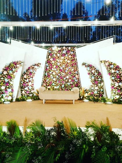 A nighttime view of a modern wedding stage, where the lighting design truly comes to life. The spotlights highlight the intricate floral work on the panels, and the hanging lights above add a magical glow.