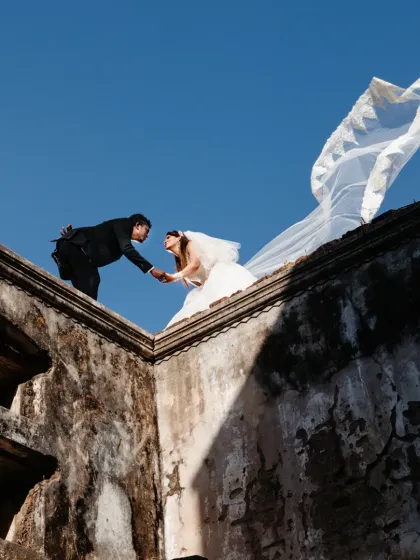 A dramatic, low-angle shot of the couple on the edge of a historic ruin. The bride's veil catching the wind against the clear blue sky creates a breathtaking moment of freedom and romance.