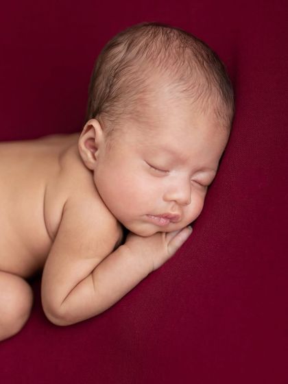 A simple, powerful portrait of a newborn sleeping on their side against a deep red backdrop. This image captures the stillness and calm of the very beginning.