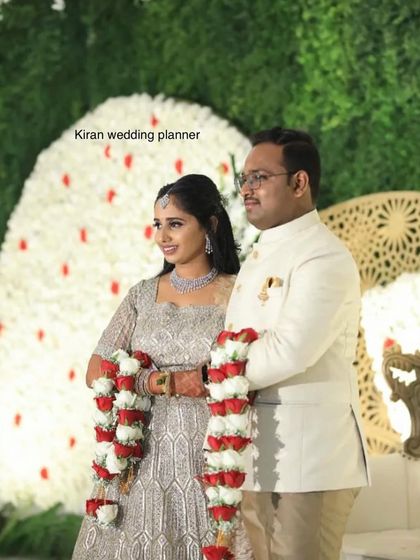 A close-up of a couple standing before a white floral wall with subtle red accents. The garlands are made of matching red and white roses, tying the whole look together.