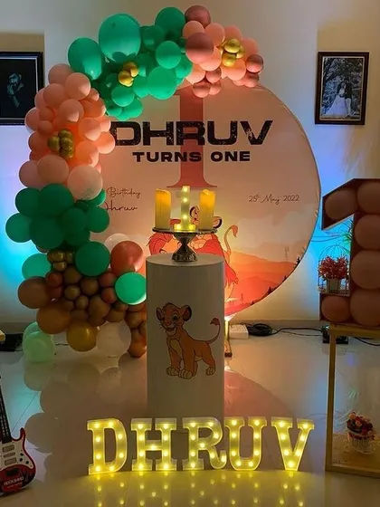 A Lion King themed first birthday party setup at home. This decoration features a custom circular backdrop, a balloon garland, a cake stand with a Simba graphic, and light-up letters spelling the name.