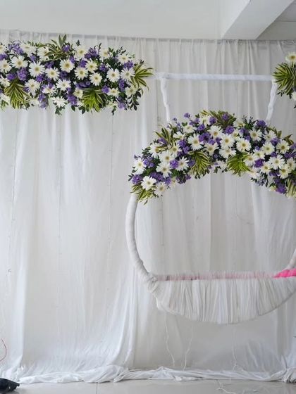 A modern and minimalist take on a naming ceremony setup. This features a simple white circular swing adorned with purple and white flowers, set against a clean white drape for a fresh and elegant look.