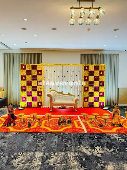 This baby shower setup uses a striking color combination of yellow and magenta in a checkerboard flower wall. The white and gold chaise lounge adds a touch of elegance, with traditional dolls and plate stands completing the scene.