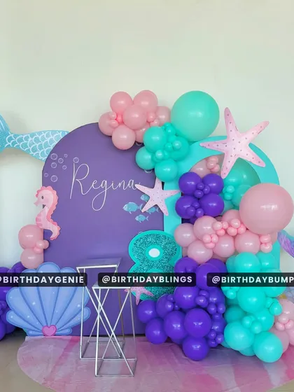 A purple and teal mermaid theme with a large shell cutout and mermaid tail accents. The balloon garland and seahorse details create a beautiful underwater photo corner.