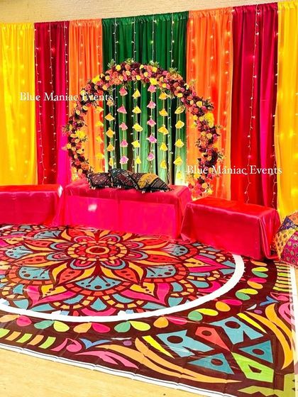 A vibrant floor-seating Mehendi setup with a colorful mandala-print carpet. The backdrop features multi-colored drapes and a circular floral arch, perfect for a bohemian-themed event.