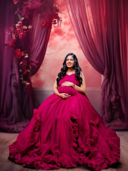 A dramatic and glamorous studio portrait. The mother-to-be is seated in a magnificent magenta tulle gown, set against a rich, curtained backdrop.