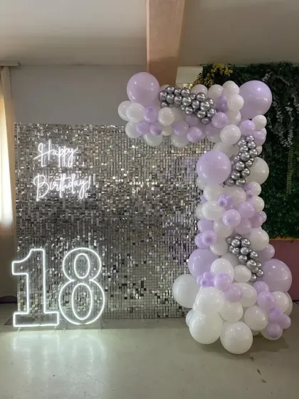 A soft lavender and white balloon arrangement for an 18th birthday, set against a silver sequin wall with neon signs.