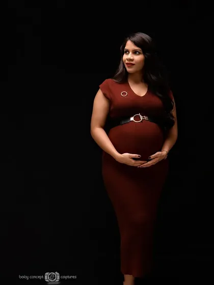 A classic and elegant low-key portrait of an expecting mother in a form-fitting maroon dress. The directional lighting beautifully sculpts her form against the black background, emphasizing the baby bump.