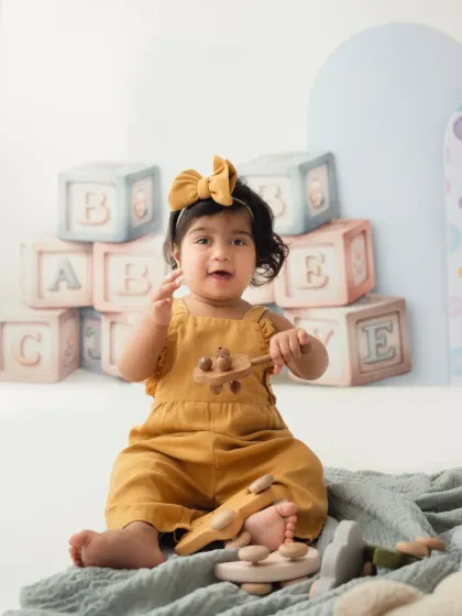 A close-up of a toddler girl in a mustard yellow romper, playing with a wooden toy car. These detail shots are a lovely part of a baby photoshoot.