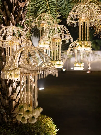 A magical detail from an outdoor event: intricate, kalire-style chandeliers hanging from a palm tree, illuminated for the evening.