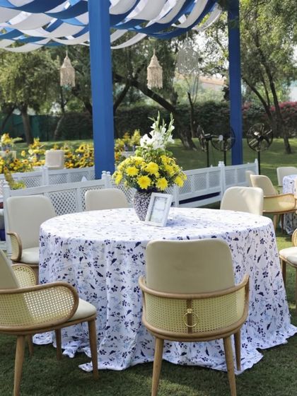 Elegant guest seating at an outdoor haldi. The round tables are covered with blue and white printed cloths, and the modern chairs with cane details add a touch of sophistication.