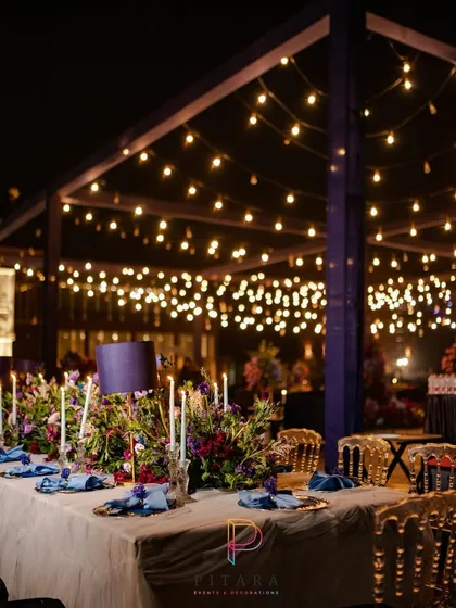 A long dining table set for a feast under the stars. The overhead structure with string lights provided a warm canopy, while lampshades and florals on the table added a touch of intimate elegance.