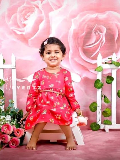 A lovely rose-themed kids' photoshoot. The little girl sits in front of a pink floral backdrop with a white picket fence, creating a sweet and classic garden party look.