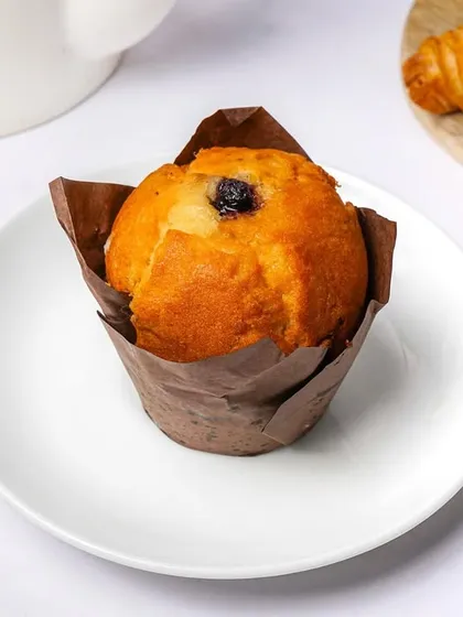 A single blueberry muffin in a brown paper liner, placed on a white plate. This simple, clean shot is perfect for a bakery's online menu or product catalog.