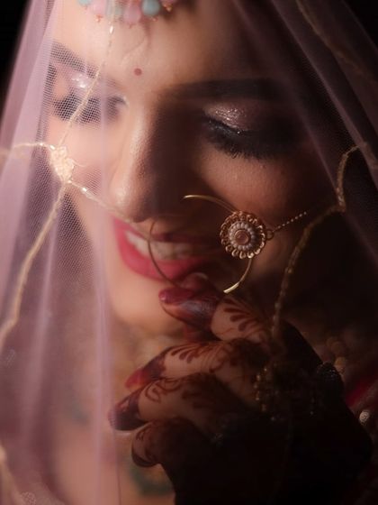 A soft, romantic shot of a bride peeking through her pink dupatta. The lighting highlights her smile and the subtle glitter on her eyes.